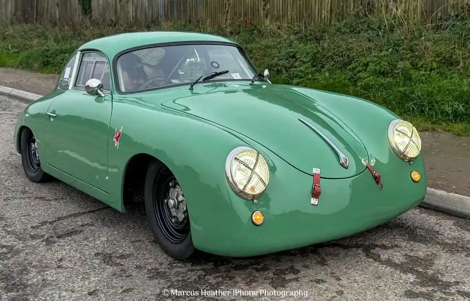 A glossy green vintage Porsche 356 sports car is parked on a paved road next to grass and a wooden fence. The car has round…
