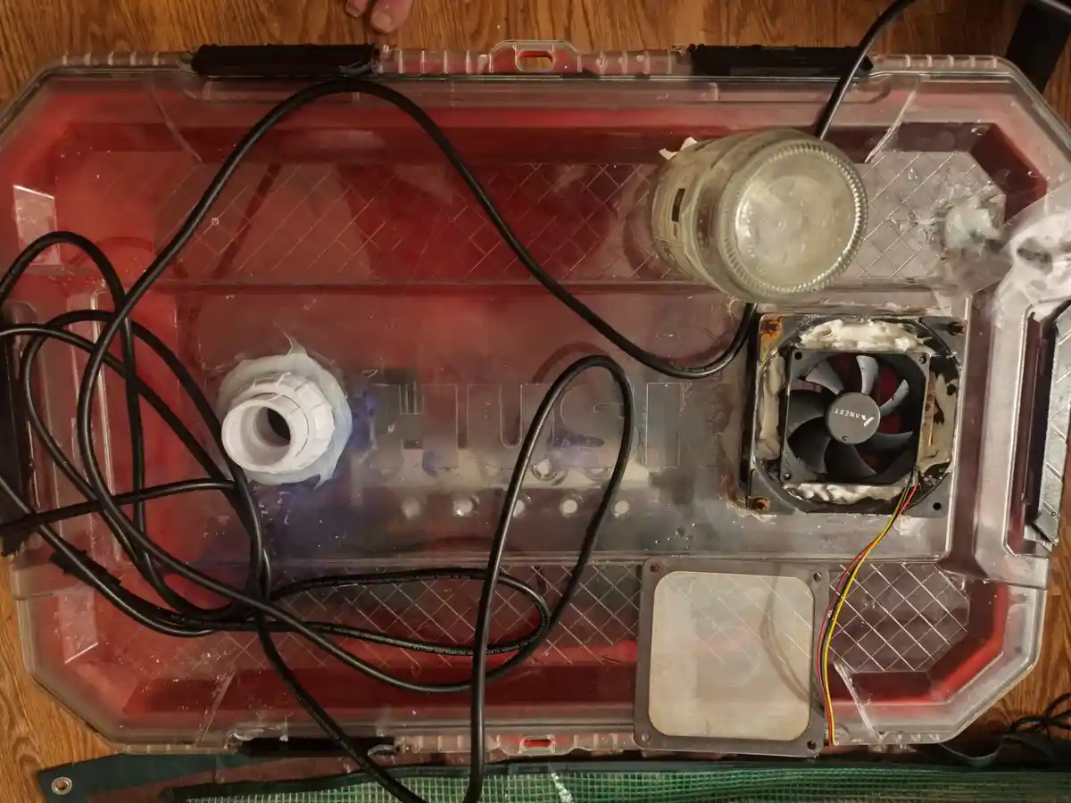A DIY computer cooling setup inside a clear plastic case filled with red liquid, featuring a fan, a glass jar, tangled black…