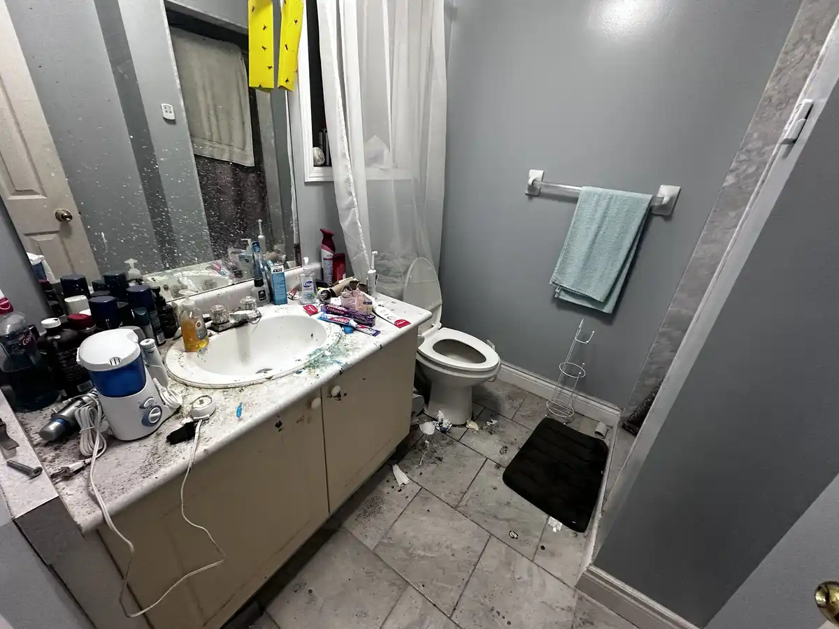 A messy bathroom with a cluttered, dirty countertop full of toiletries, a stained sink, and trash on the floor near the…
