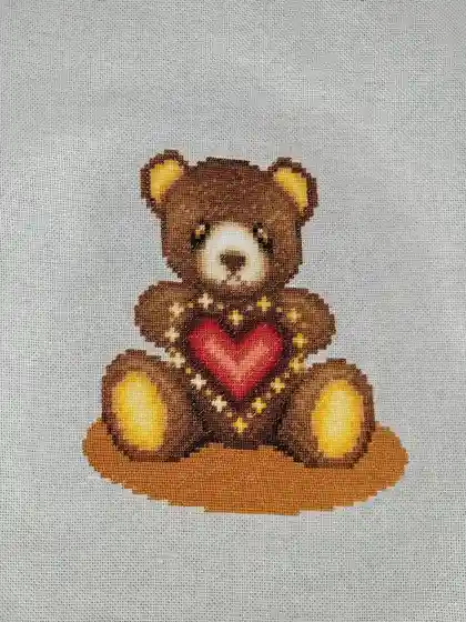 A cross-stitch pattern of a brown teddy bear sitting upright, holding a red heart outlined with yellow stars, stitched on…