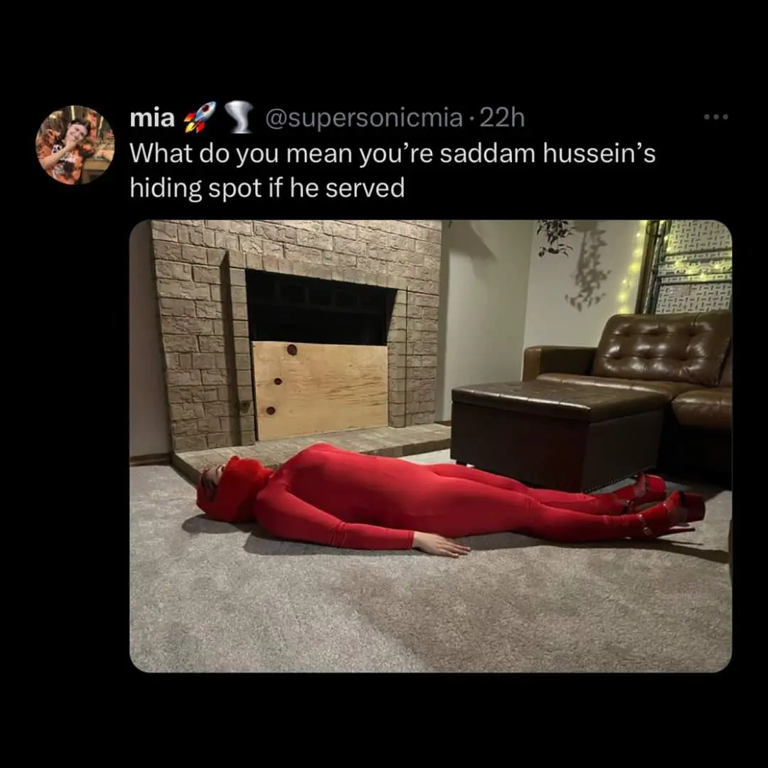A person in a full red bodysuit and red high heels lies motionless on a carpeted floor in front of a fireplace with a wooden…