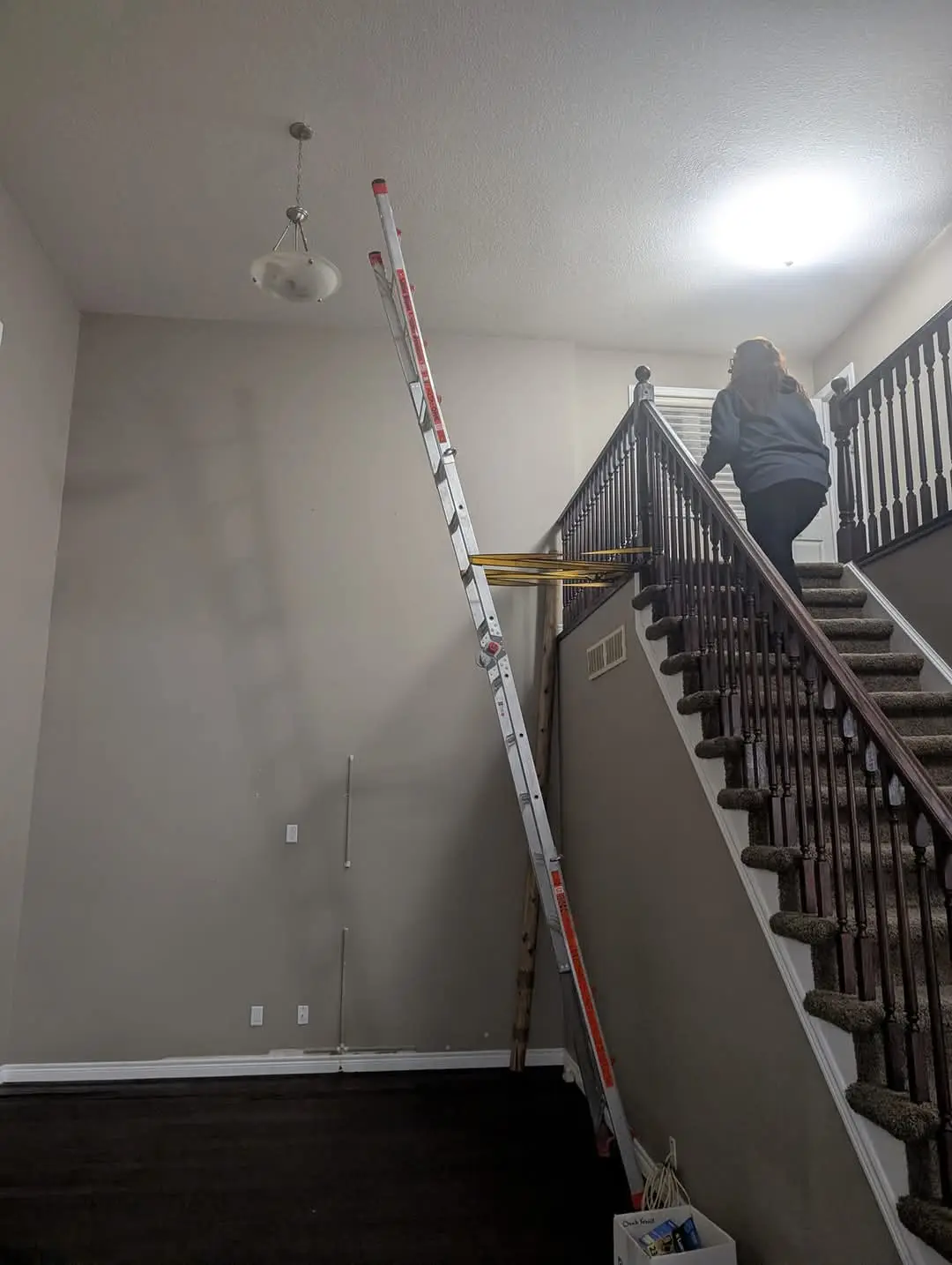 A person in a blue hoodie walks up a carpeted staircase while a tall aluminum ladder leans against the wall, reaching toward…
