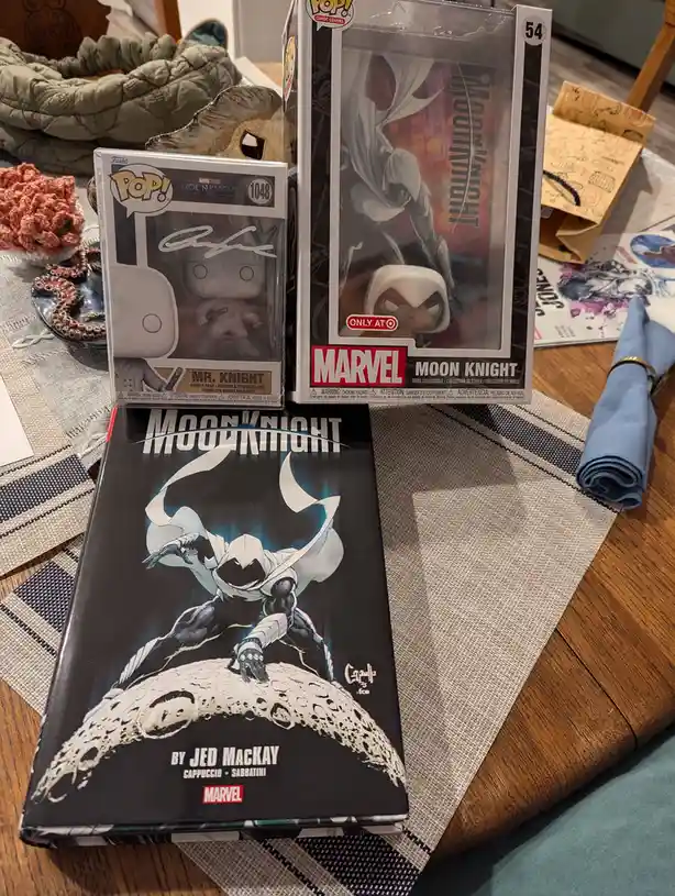 A collection of Moon Knight merchandise including a Funko Pop! vinyl figure, a larger Moon Knight comic book, and a Funko…