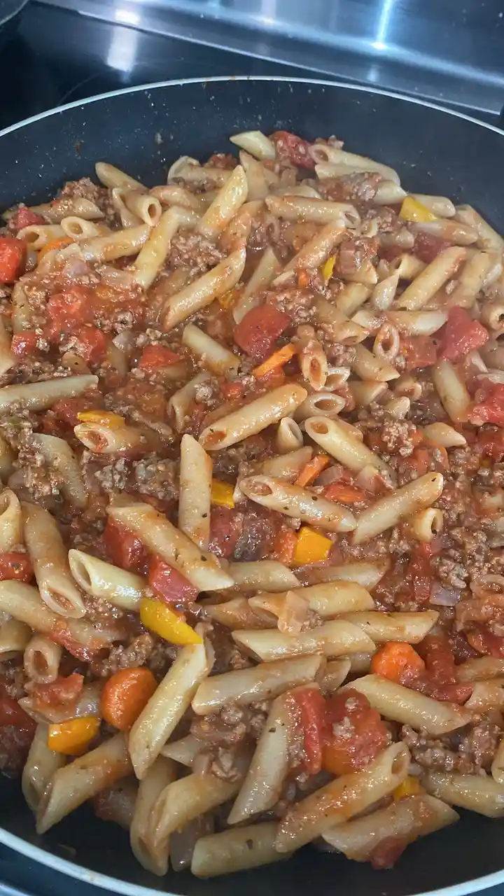 A close-up photo shows penne pasta mixed with a meat sauce, diced tomatoes, and colorful bell peppers in a black frying pan…