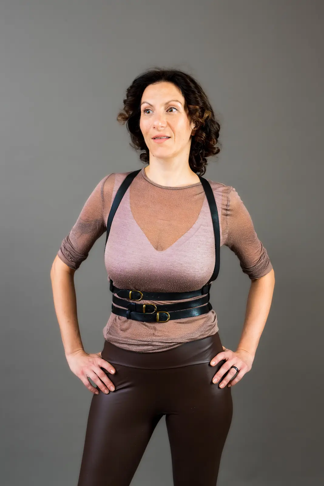 A woman with curly dark hair stands against a plain gray background, wearing a sheer brown top, brown shiny pants, and…