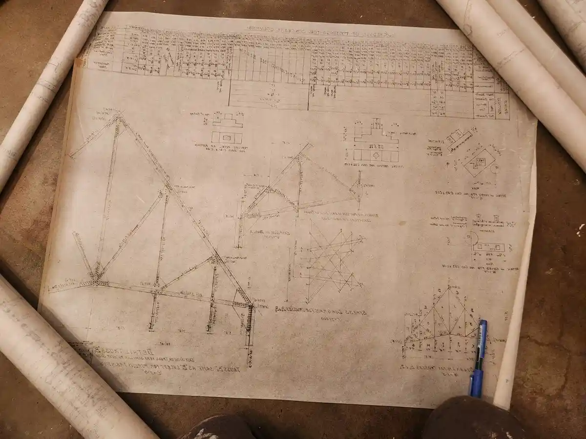An overhead shot of a large, aged engineering blueprint spread on a work surface, surrounded by rolled-up plans and a blue…