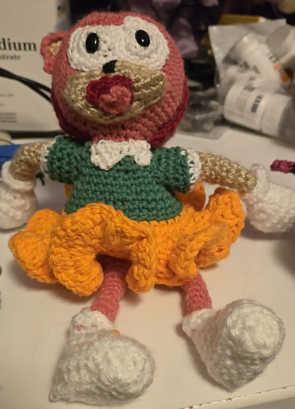 A handmade crocheted monkey doll with a pink head, tan face, red nose, green sweater with white collar, and orange skirt…