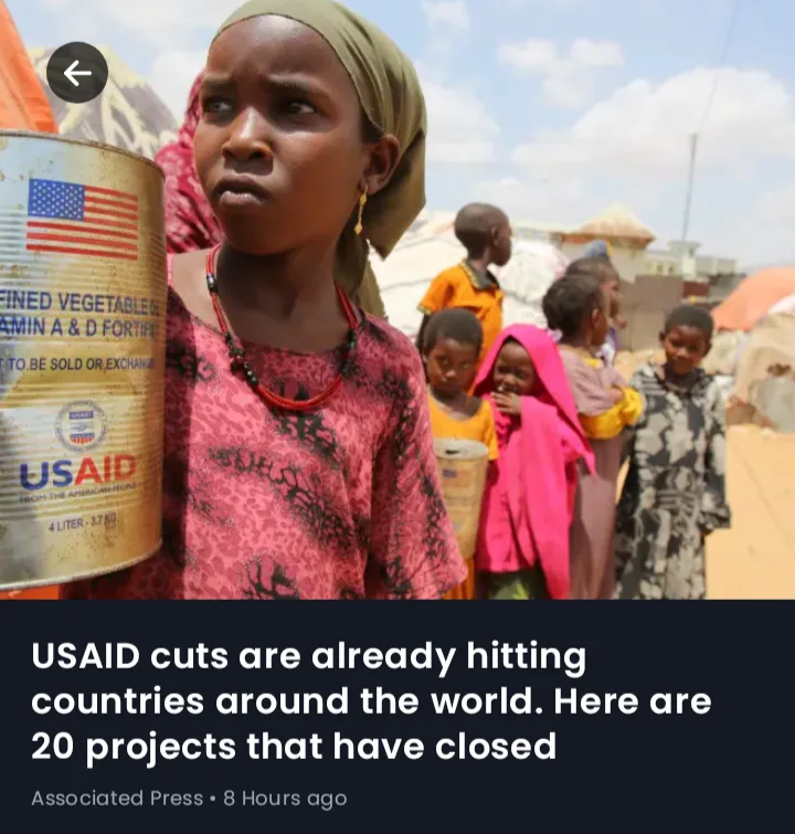 A young girl in a pink patterned shirt and green headscarf holds a large can labeled 'USAID' with an American flag, standing…