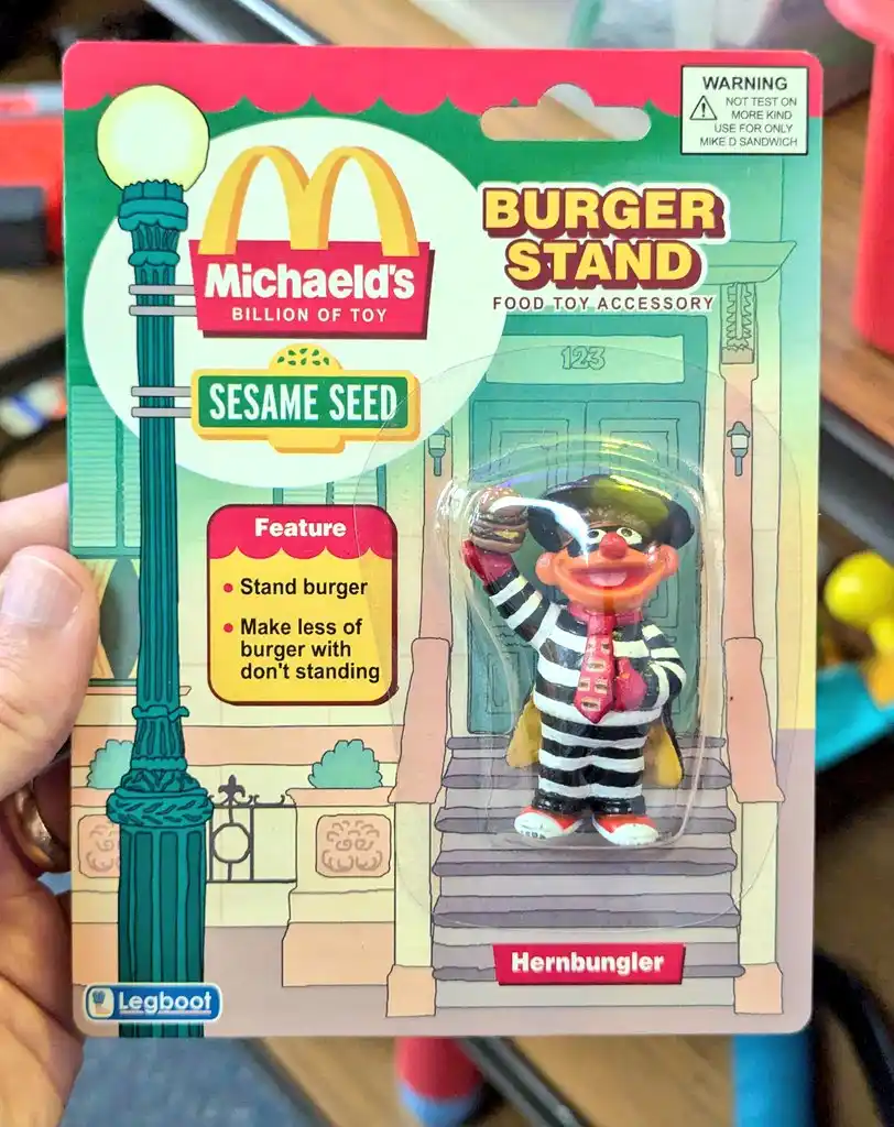 A toy package for a Sesame Street character, 'Hernbungler', dressed in a striped outfit holding a burger, labeled as a…
