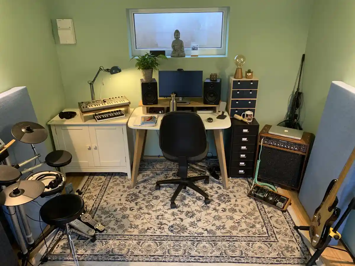 A cozy home studio room with light green walls, a patterned rug, and a desk with a computer, speakers, and a microphone. To…