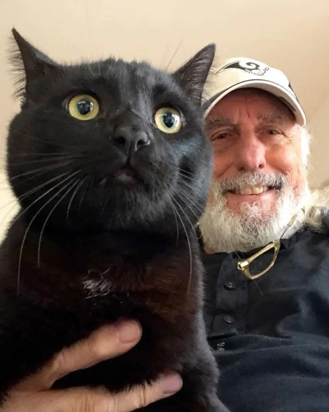 A man with a white beard and a baseball cap is smiling while holding a black cat with wide, startled-looking yellow eyes…