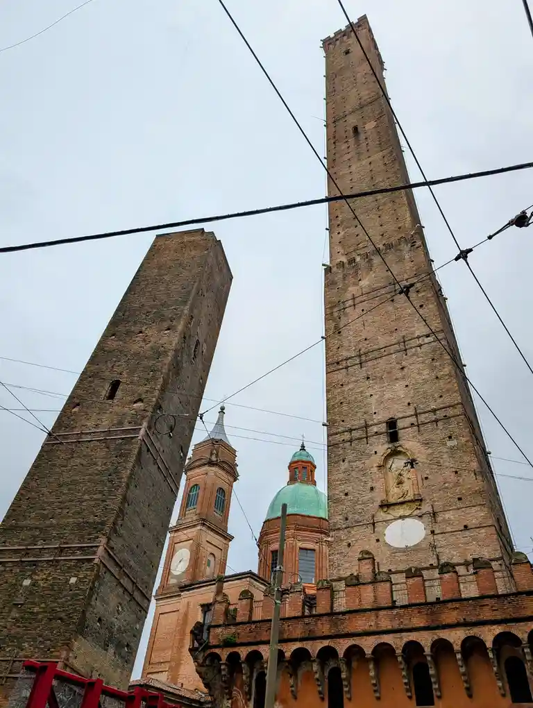 A low-angle photo shows two tall, ancient brick towers leaning toward each other, appearing to be in a standoff. Between…