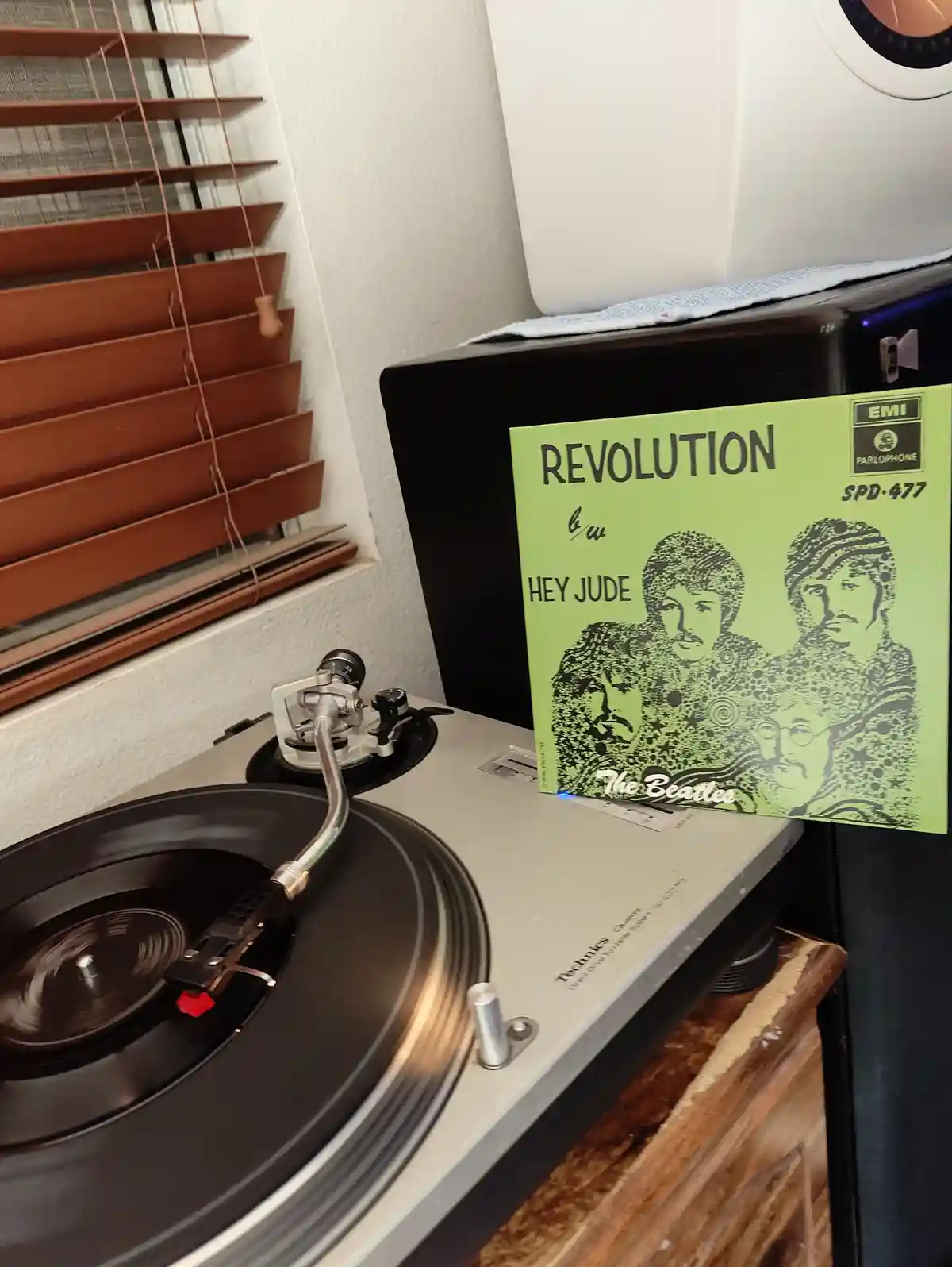 A Technics turntable spins a record while a green Beatles album cover titled 'Revolution' leans against it, featuring the…