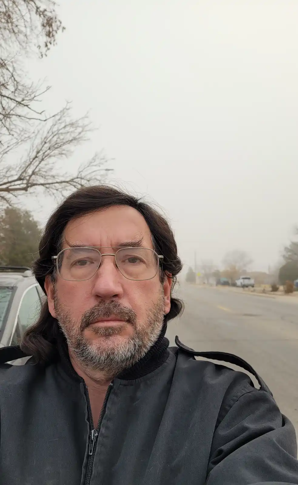A man with long dark hair, glasses, and a gray beard takes a selfie outdoors on a hazy day. Behind him is a street with cars…