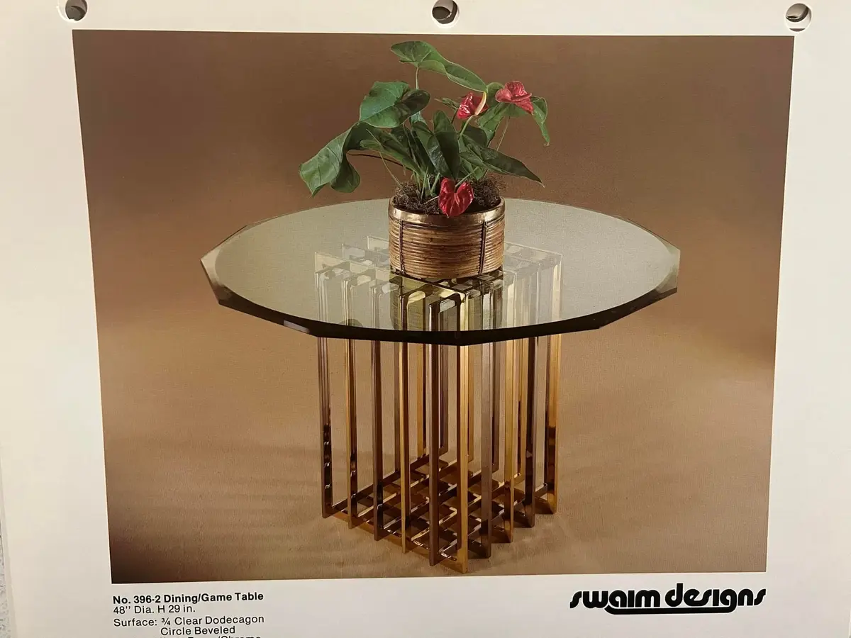 A vintage catalog photo shows a glass-topped table with a brass-colored, geometrically patterned base, holding a potted…