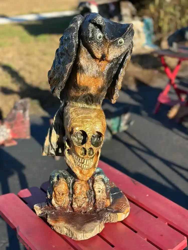 A carved wooden sculpture depicts an owl perched atop a human skull, which rests on a base with three smaller carved skulls…