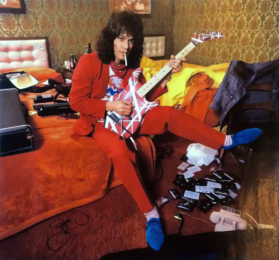 A man with long dark hair, wearing a red suit and blue socks, sits on a bed holding a red and white electric guitar with a…
