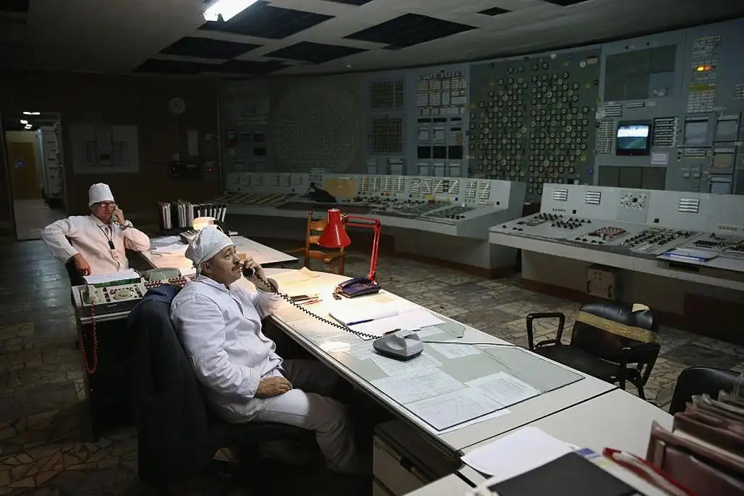 Two men in white uniforms and caps sit at desks in a dimly lit control room filled with panels, dials, and vintage…