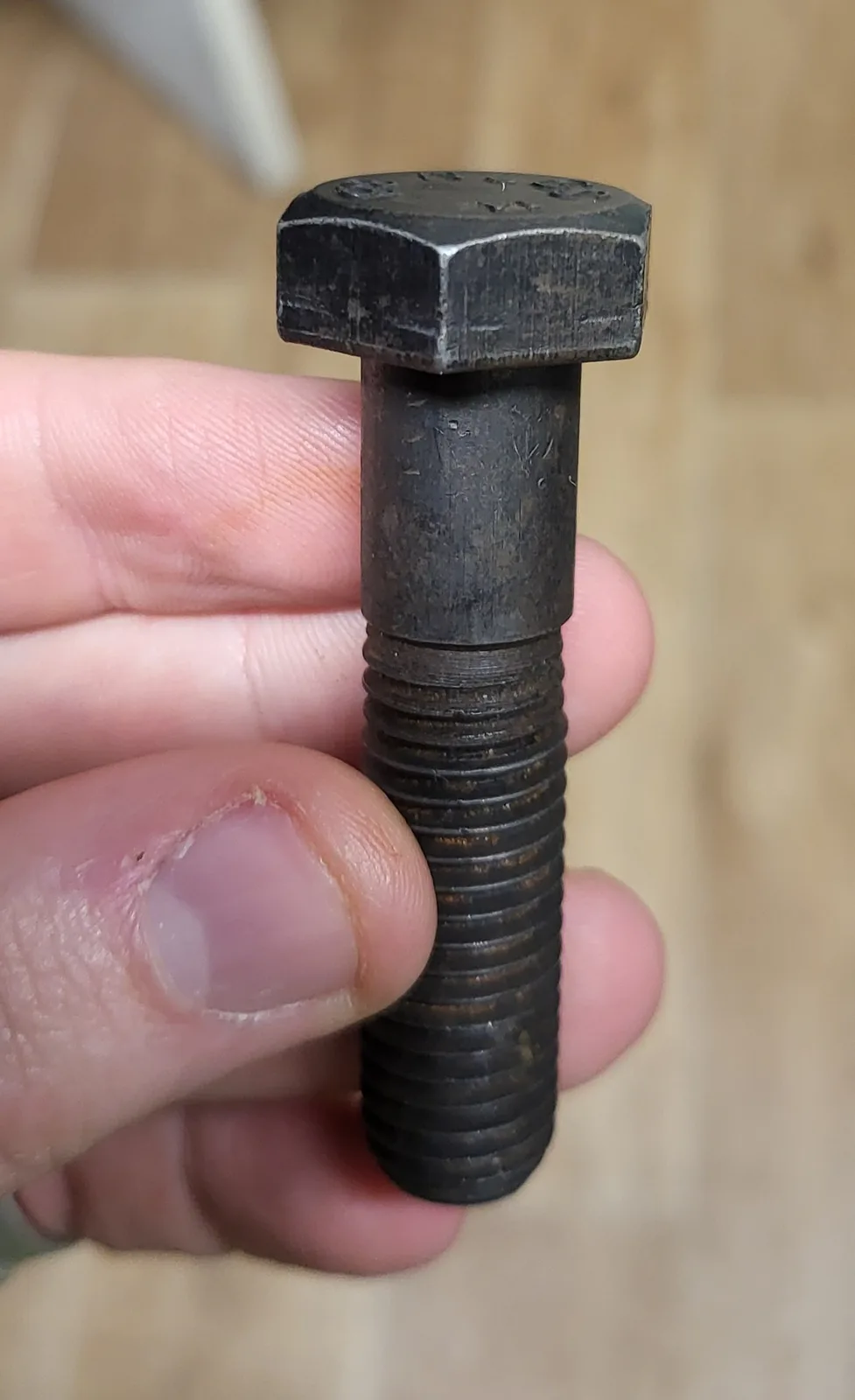 A close-up photo shows a person's hand holding a large, dark, rusty hex bolt with visible threading. The bolt appears worn…