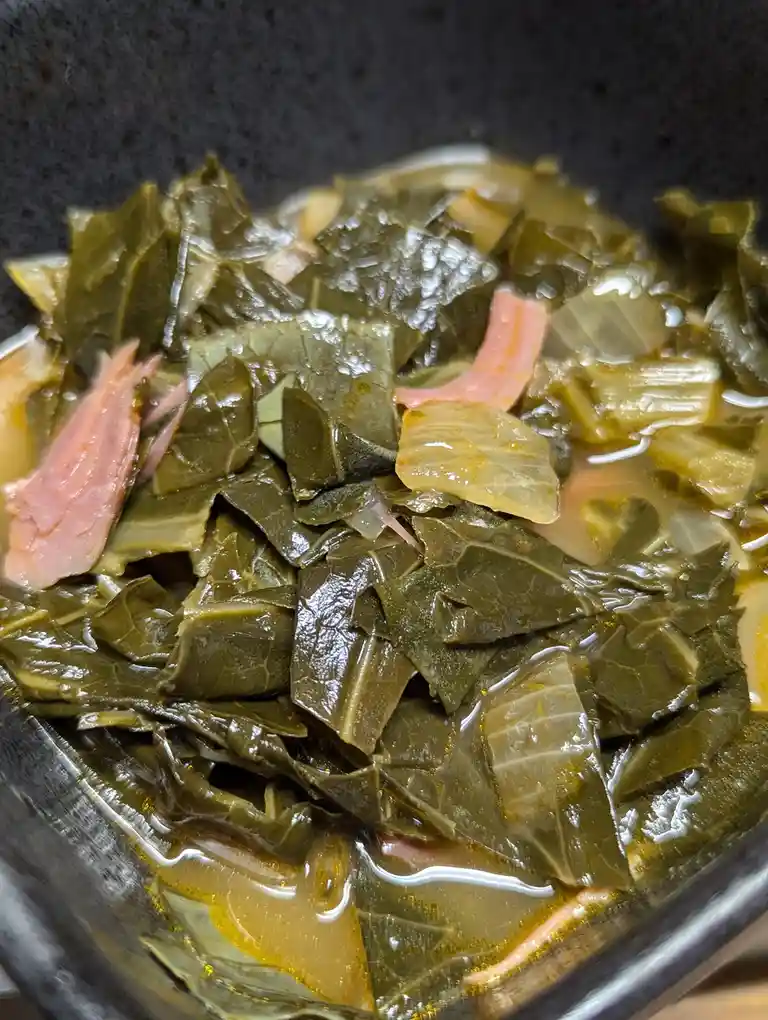 A close-up photo shows a pot of cooked collard greens with pieces of ham and onions in a brownish broth. The greens are dark…