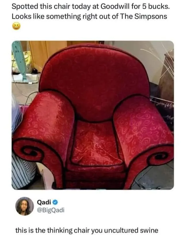 A red, patterned armchair with exaggerated, cartoonishly curved arms and a rounded back is shown in a cluttered room…