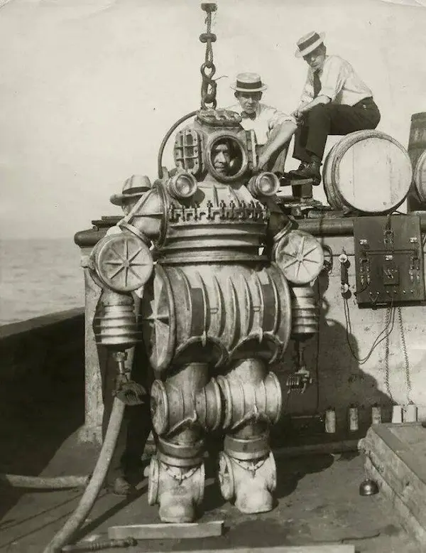 A vintage black-and-white photograph shows a large, bulky, 19th-century diving suit standing on a ship's deck, with two men…