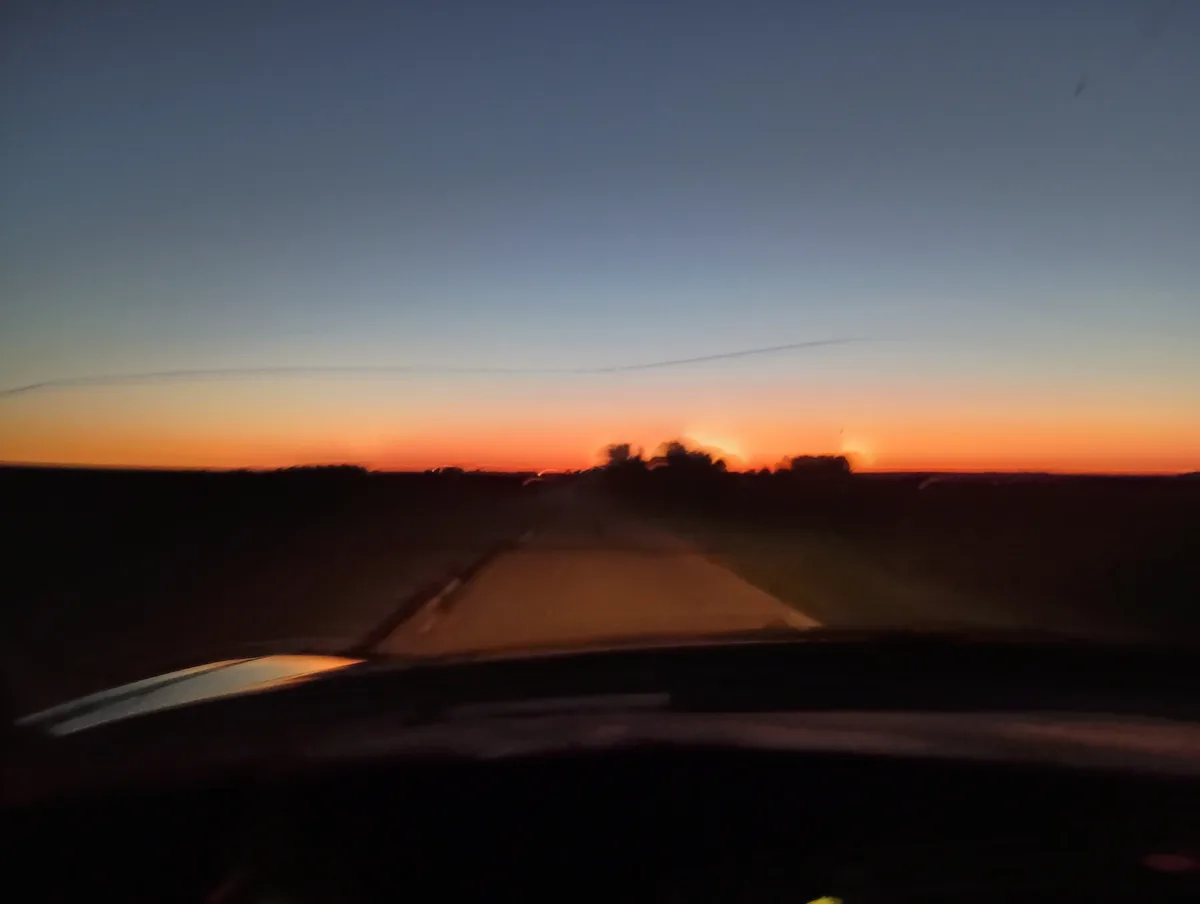 A blurry photo taken from inside a moving car at sunset, showing a road ahead and a vibrant orange and blue sky. The…
