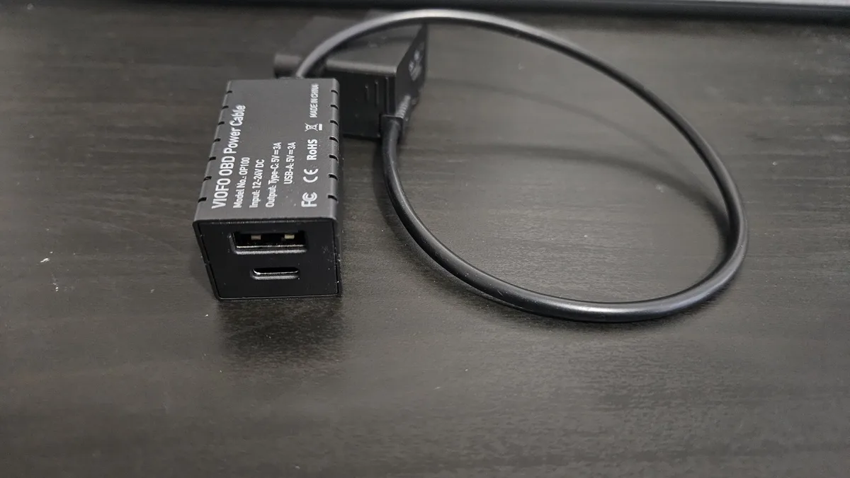 A black VIOFO OBD Power Cable with a USB-A and USB-C port sits on a dark surface, its cable looped into a circle. The text…
