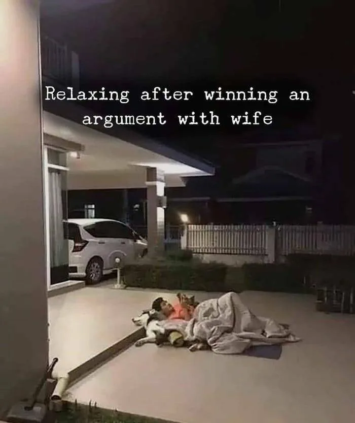 A man lies on a mat outside a house at night, covered by a blanket, with two dogs resting beside him. A white car is parked…