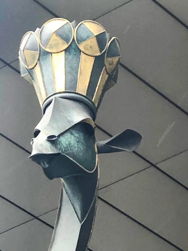 A close-up of a stylized, metallic sculpture of a bird's head, possibly a crane or heron, wearing a chef's hat with gold and…