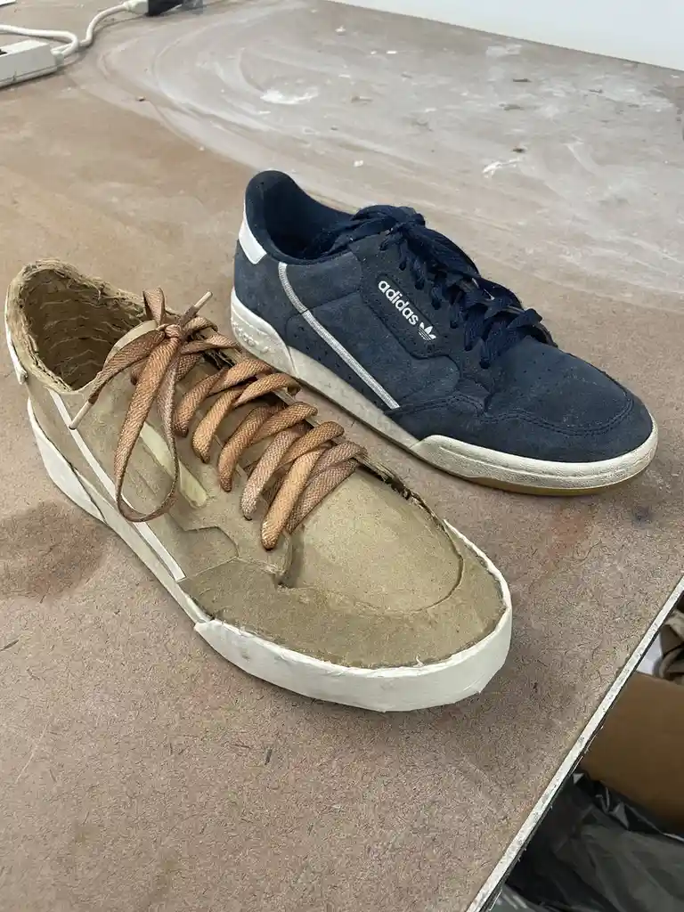 Two sneakers sit side by side on a worn wooden surface; one is a real navy blue Adidas sneaker with white stripes and laces…