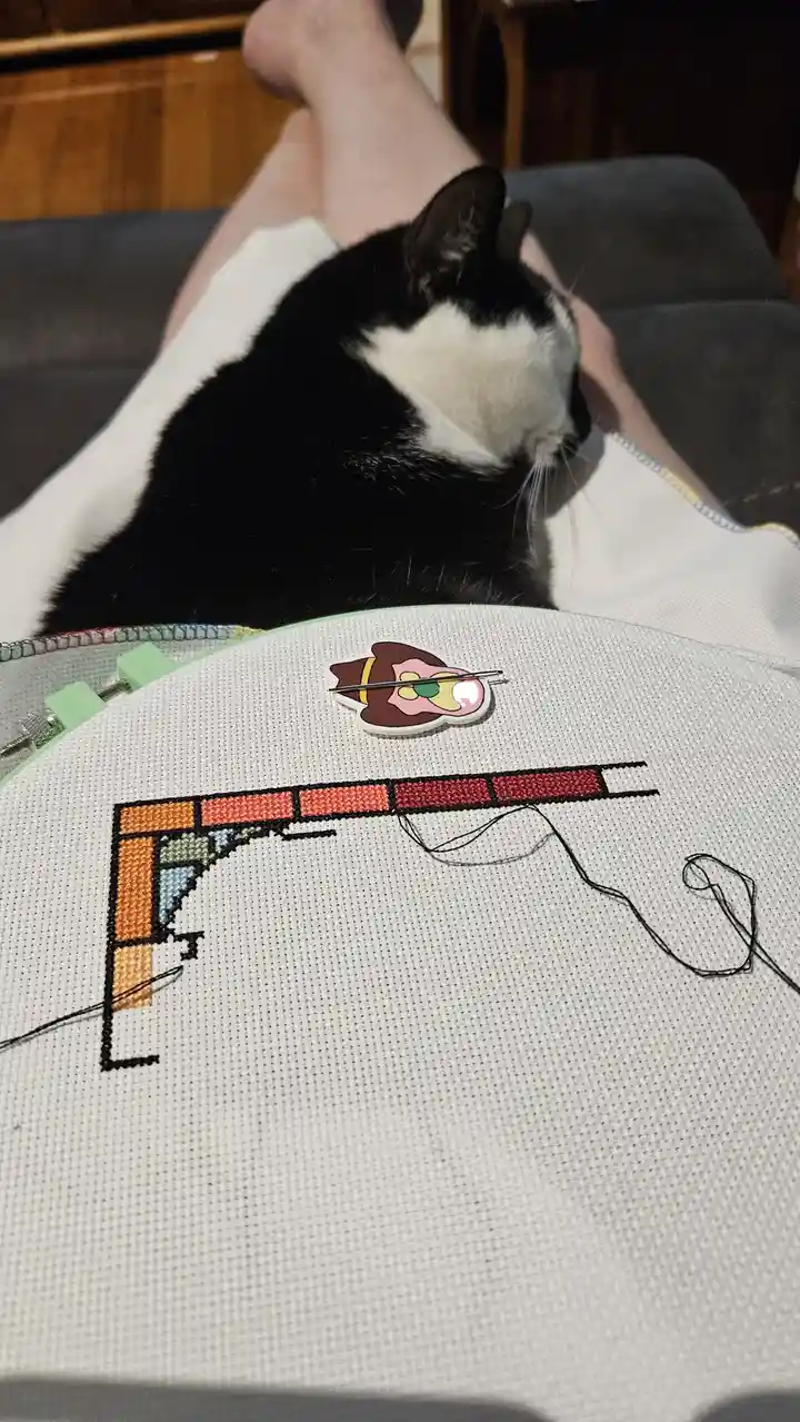 A black and white cat is curled up asleep on a person's lap, which is holding an embroidery hoop. The hoop contains a…