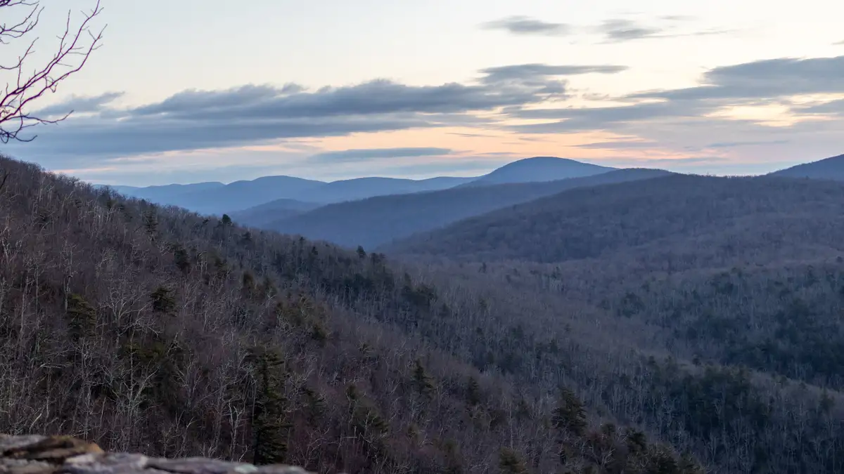 A serene landscape photo of rolling, forested mountains under a cloudy twilight sky, with bare trees and distant ridges…