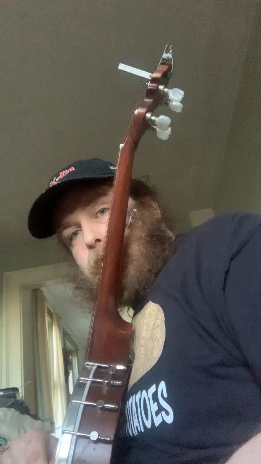 A man with a long beard and a black cap holds a banjo vertically, obscuring most of his face except his eyes and nose. He…