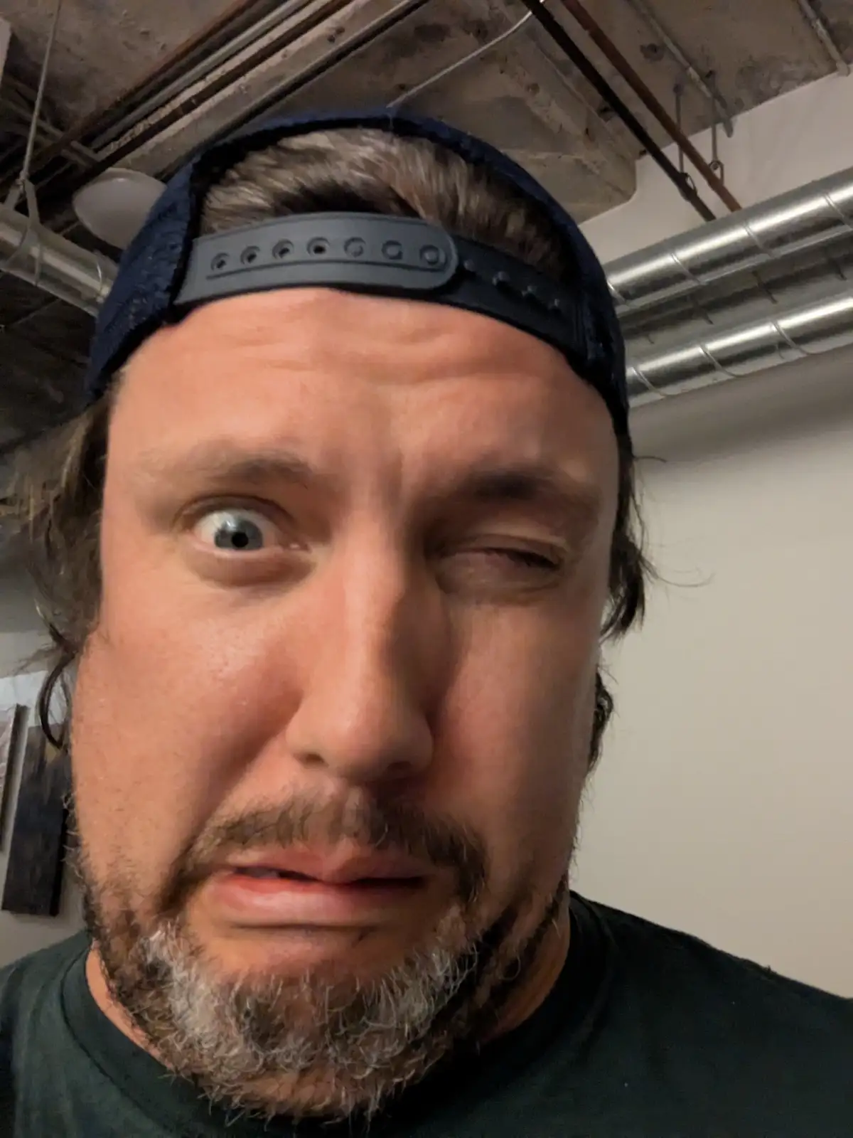 A man with a beard and a backwards baseball cap stares intensely at the camera with one eye wide open and the other squinted…