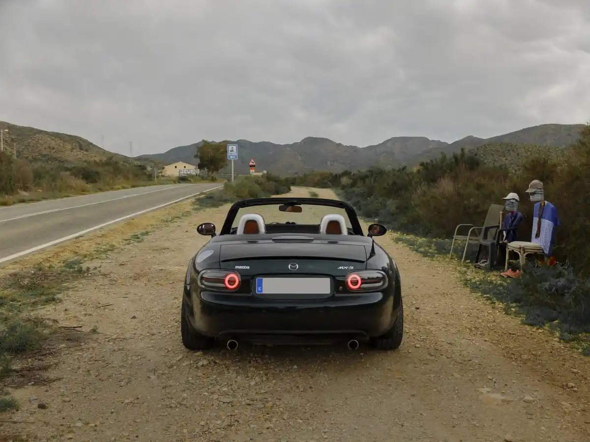 A black Mazda MX-5 Miata convertible is parked on a gravel shoulder next to a paved road, facing away from the camera. On…