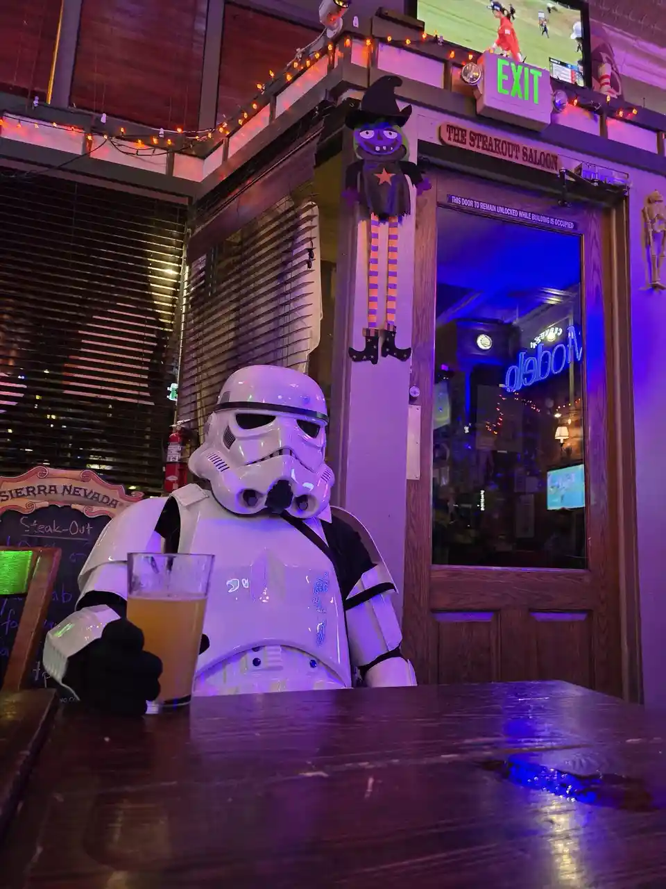 A person in a full white stormtrooper costume sits at a wooden bar in a dimly lit, purple-lit bar called 'The Steakout…