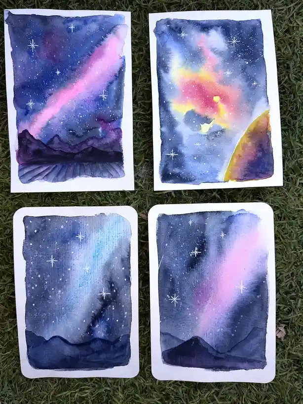 Four watercolor paintings of starry night skies with colorful auroras and mountain silhouettes lie on green grass. Each…