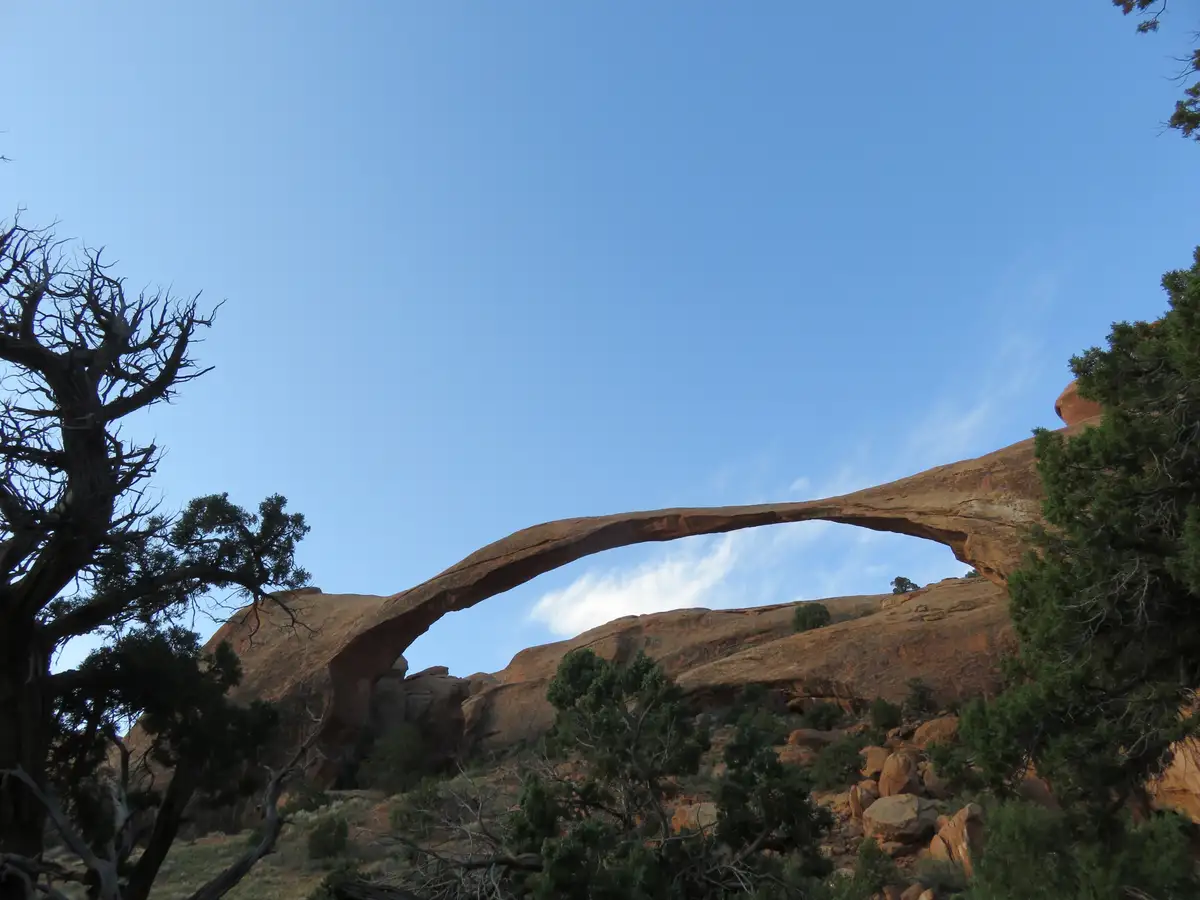A low-angle photo shows a large, natural sandstone arch spanning across a desert landscape under a clear blue sky, framed by…
