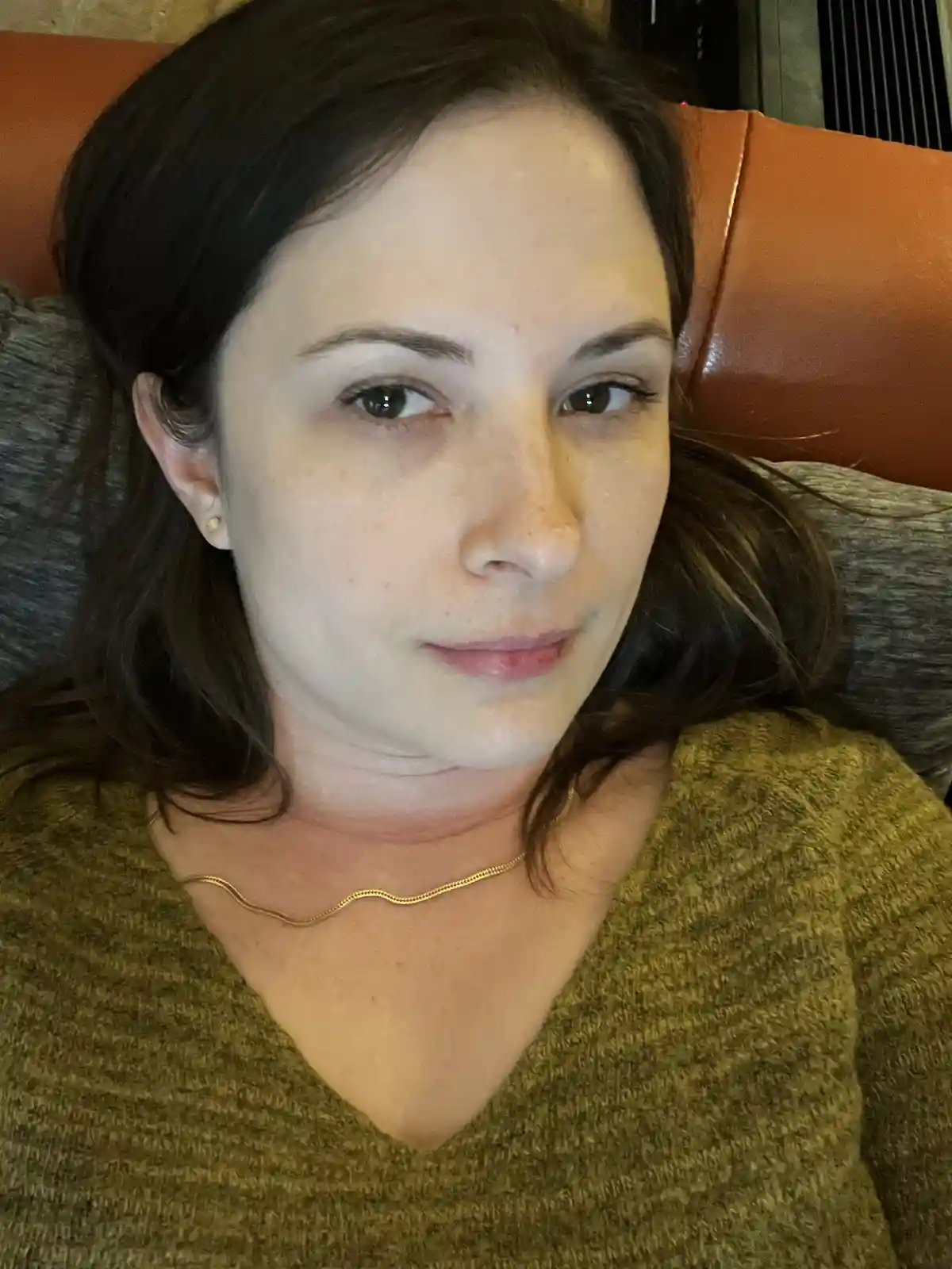 A woman with dark hair and a gold chain necklace looks directly at the camera with a neutral expression while lying on a…