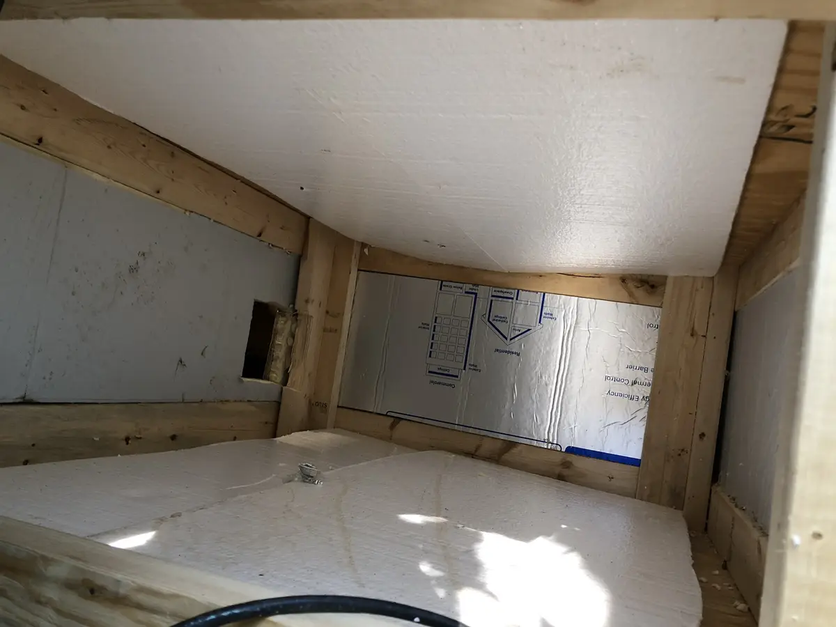 A low-angle photo shows the unfinished interior of a small attic or crawlspace with exposed wooden beams, white insulation…