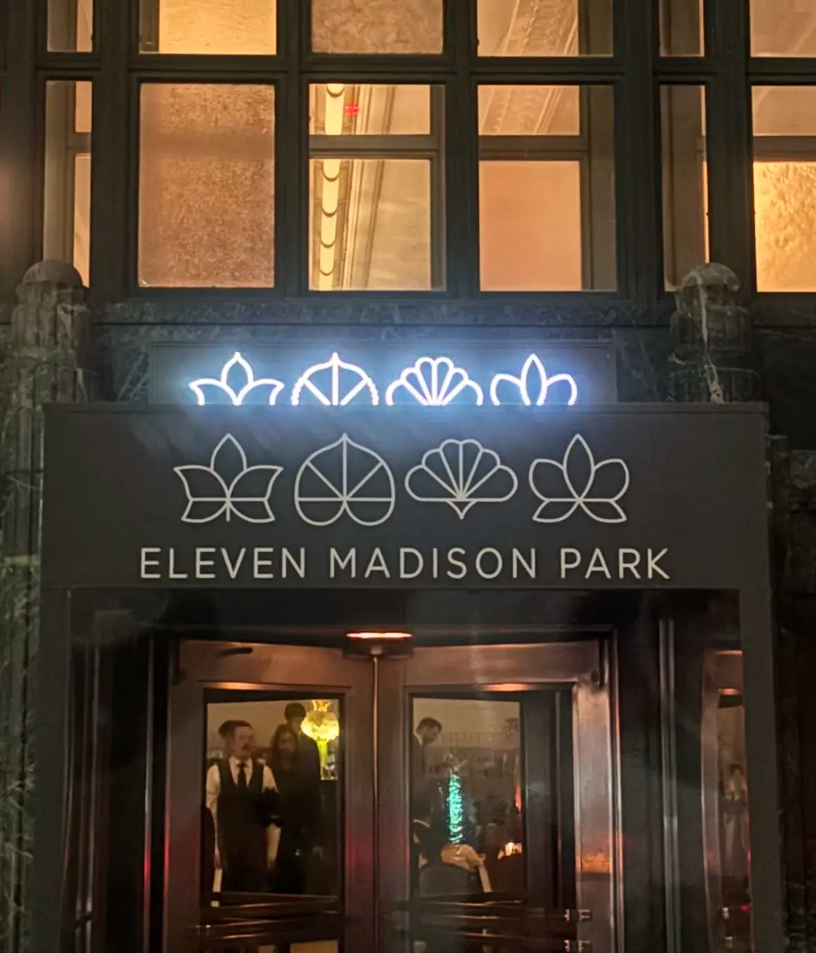 A nighttime photo of the entrance to the upscale restaurant Eleven Madison Park, featuring its illuminated sign with…