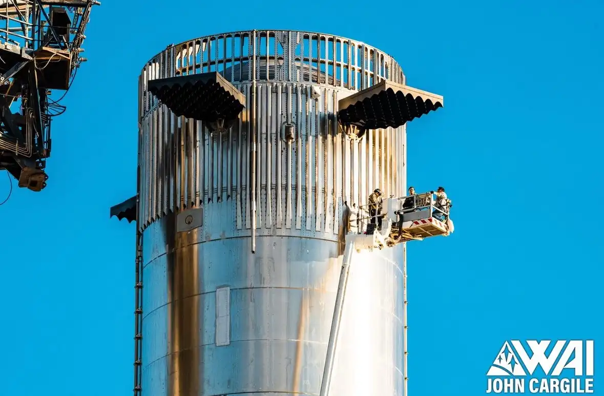 A photo shows three workers in a cherry picker on the side of a tall, cylindrical metal structure against a clear blue sky…