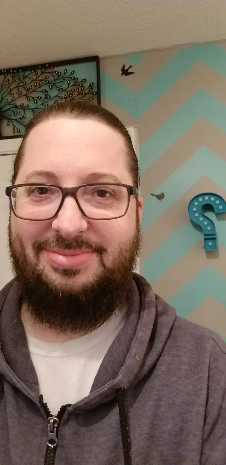 A man with glasses and a beard smiles at the camera while standing in front of a wall with a teal and beige chevron pattern…