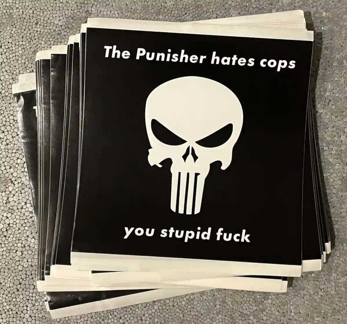 A stack of black square stickers with a white Punisher skull logo, overlaid with the text 'The Punisher hates cops' at the…