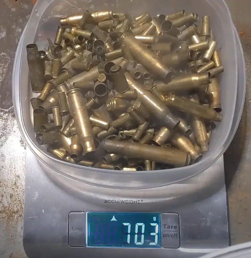 A clear plastic container filled with brass bullet casings sits on a digital AccuWeight scale, which displays the weight as…