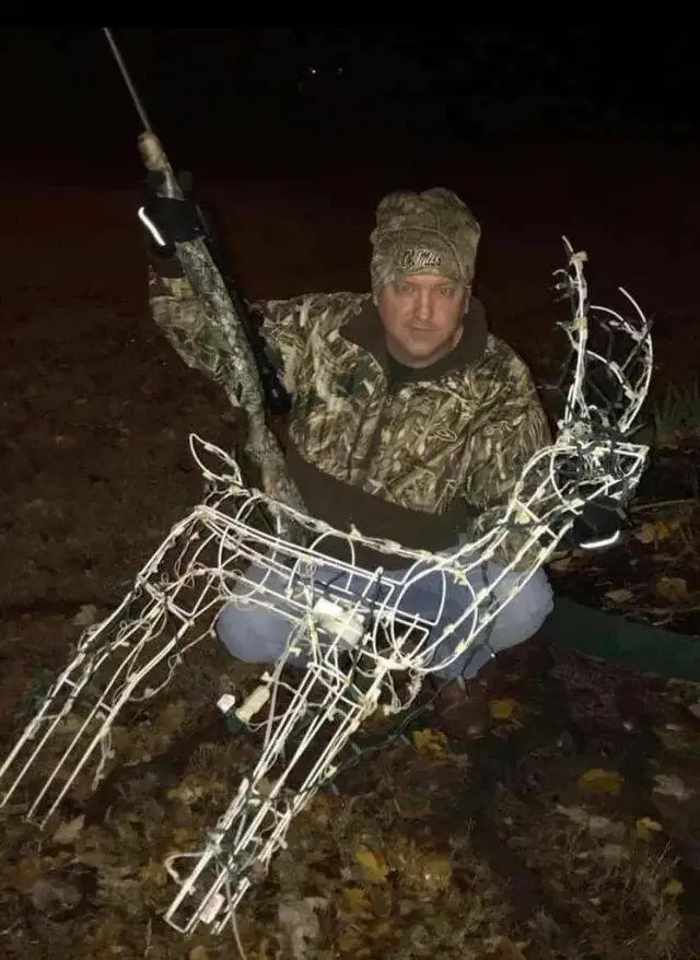 A man in camouflage clothing and a beanie kneels outdoors at night, holding a rifle aloft in one hand while posing beside a…