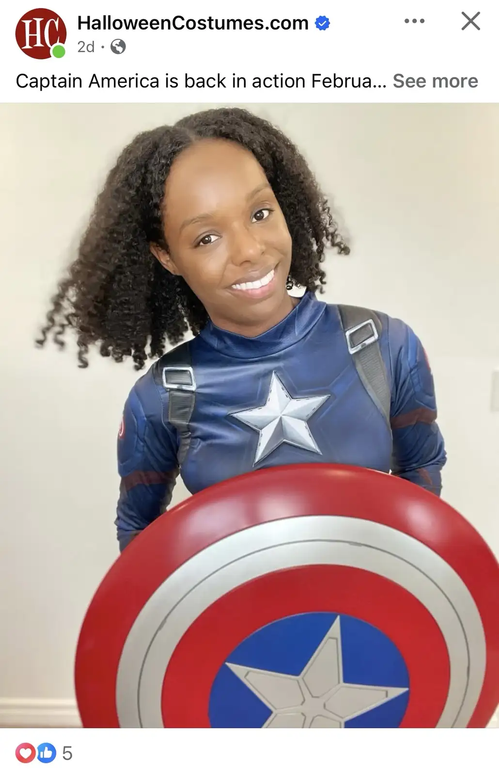 A woman with curly dark hair is smiling while wearing a blue Captain America costume with a silver star on the chest and…