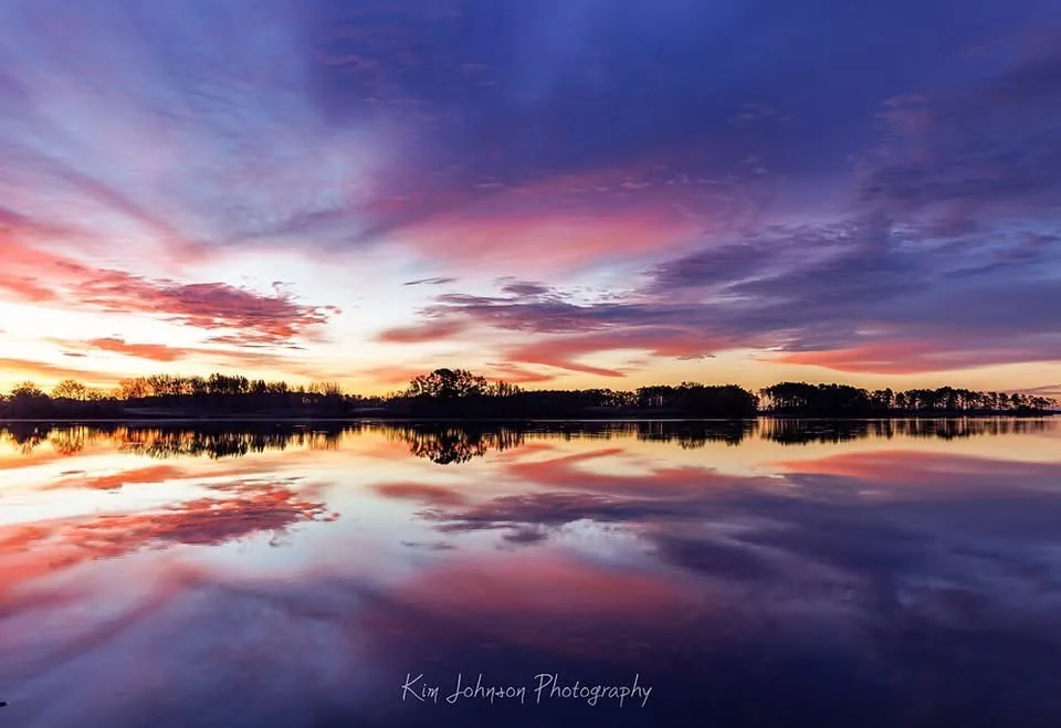 A serene landscape photograph of a calm body of water reflecting a vibrant sunset sky filled with pink, orange, and purple…