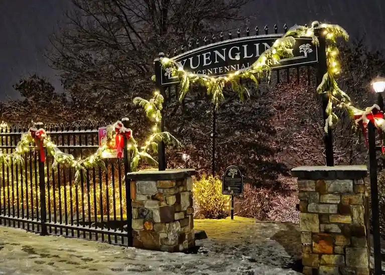 A nighttime photo of a snow-dusted park entrance with a black wrought iron gate and stone pillars, decorated with festive…